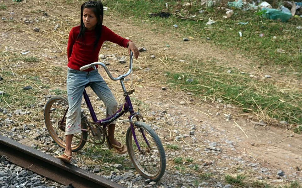 Ein Mädchen in rotem Pullover und Jeans steht auf einem lila Fahrrad neben einem Eisenbahngleis, im Hintergrund Gras und Müll.