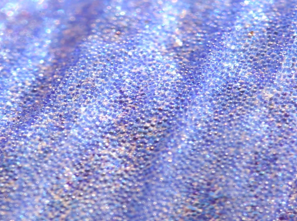 Nahaufnahme einer blauen, körnig strukturierten Oberfläche mit vielen kleinen Noppen und hellen Glanzpunkten.