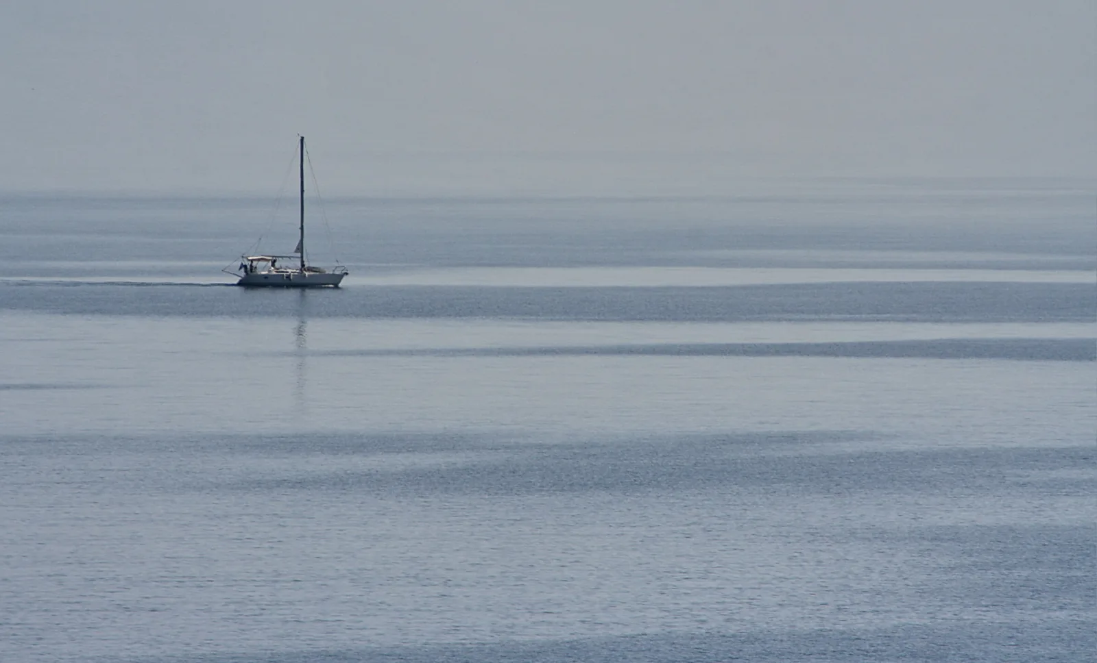 Ein kleines weißes Segelboot mit hohem Mast fährt auf einem ruhigen, graublauen Meer unter diesigem Himmel.