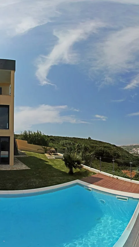 Türkisblaues Schwimmbecken vor einem gelben Haus mit Glasgeländer, dahinter grüne Hügel und blauer Himmel mit Schleierwolken.