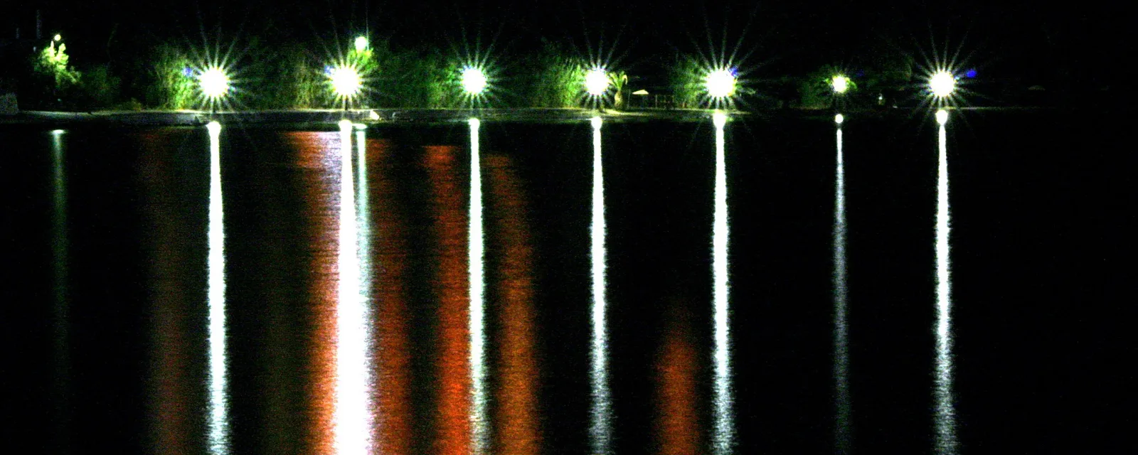 Eine Reihe beleuchteter Straßenlaternen am Ufer, deren Licht sich nachts im dunklen Wasser spiegelt.