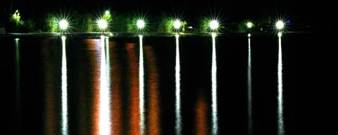 Eine Reihe beleuchteter Straßenlaternen am Ufer, deren Licht sich nachts im dunklen Wasser spiegelt.