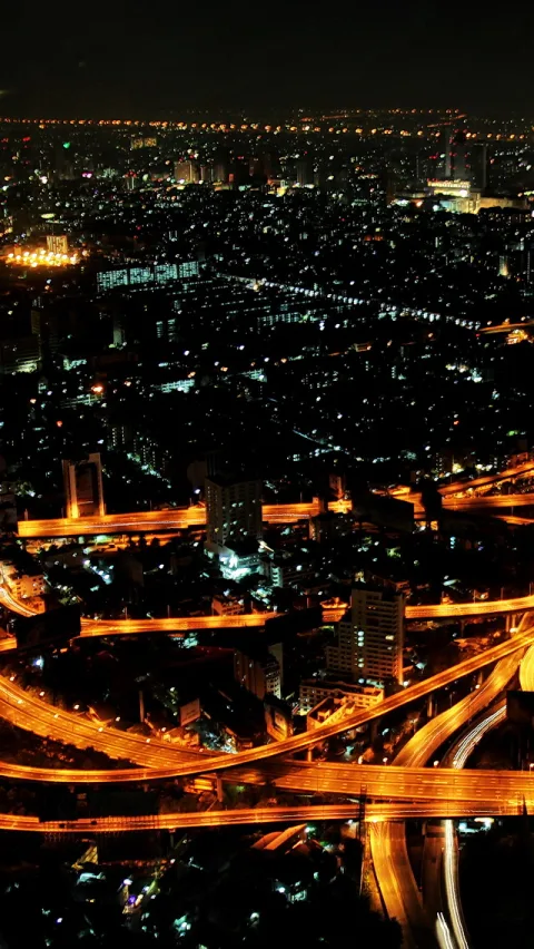 Luftaufnahme einer Großstadt bei Nacht mit mehrspurigen, orange beleuchteten Hochstraßen, die sich kreuzen und Lichtspuren hinterlassen.