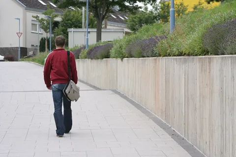 Ein Mann in roter Jacke und Jeans geht von hinten einen hell gepflasterten Gehweg entlang, eine Umhängetasche über der Schulter. Rechts steht eine Betonmauer mit blühendem Lavendel, im Hintergrund Häuser und Laternen.