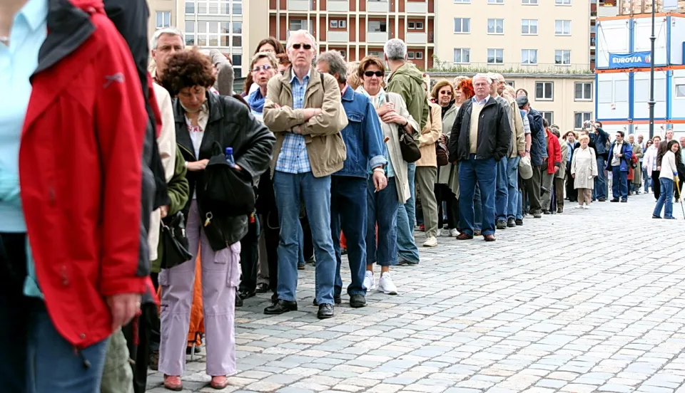 Eine lange Warteschlange von Menschen in Jacken und Hemden steht auf einem Kopfsteinpflaster vor einer Stadtbebauung, im Hintergrund ein Baugerüst mit blauer Werbung.