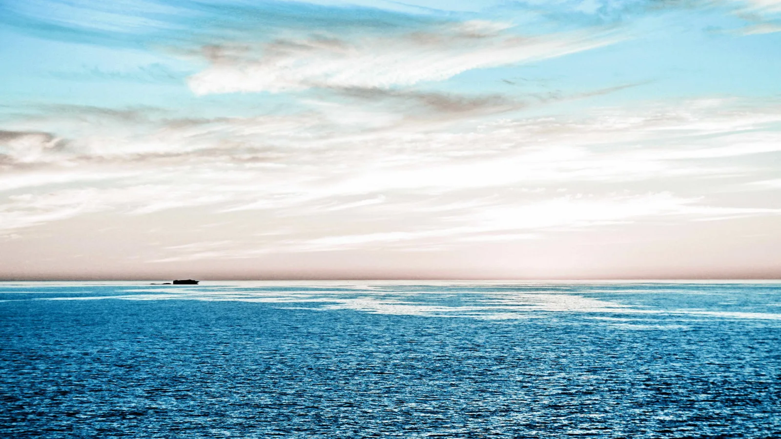 Ruhiges blaues Meer unter einem pastellfarbenen Himmel mit feinen Wolkenstreifen, am Horizont liegt eine kleine dunkle Insel.