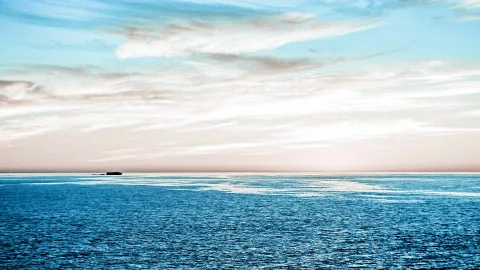 Ruhiges blaues Meer unter einem pastellfarbenen Himmel mit feinen Wolkenstreifen, am Horizont liegt eine kleine dunkle Insel.