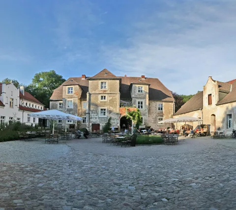 Innenhof eines alten Herrenhauses mit Kopfsteinpflaster, im Vordergrund Cafétische mit Stühlen und weißen Sonnenschirmen, dahinter die verputzte Fassade mit Rundbogeneinfahrt unter blauem Himmel.