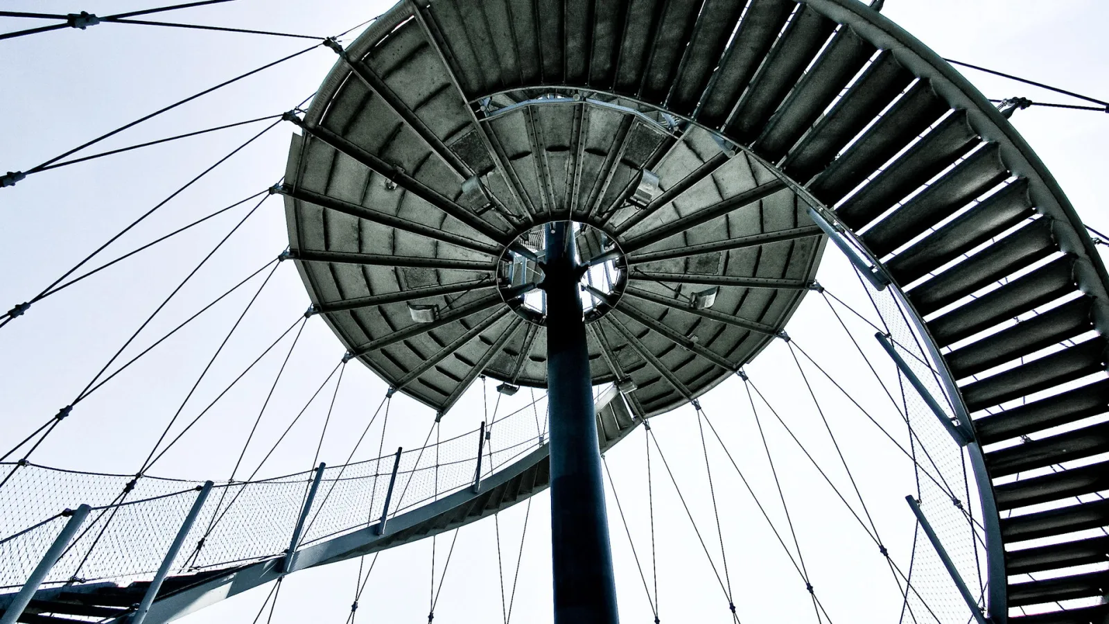 Blick von unten auf einen Stahlturm mit spiralförmiger Treppe und gespannten Stahlseilen vor hellem Himmel.