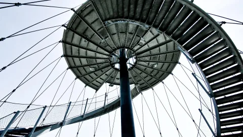 Blick von unten auf einen Stahlturm mit spiralförmiger Treppe und gespannten Stahlseilen vor hellem Himmel.