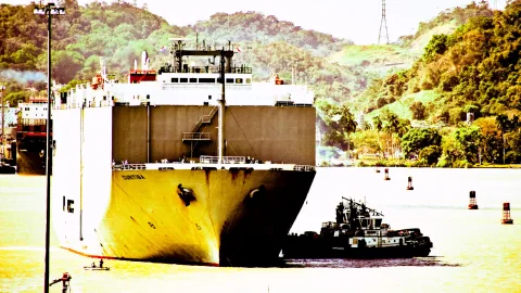Gelbes Frachtschiff mit hohem Aufbau fährt durch den Panama-Kanal, davor ein kleiner Schlepper, im Hintergrund bewaldete Hügel.