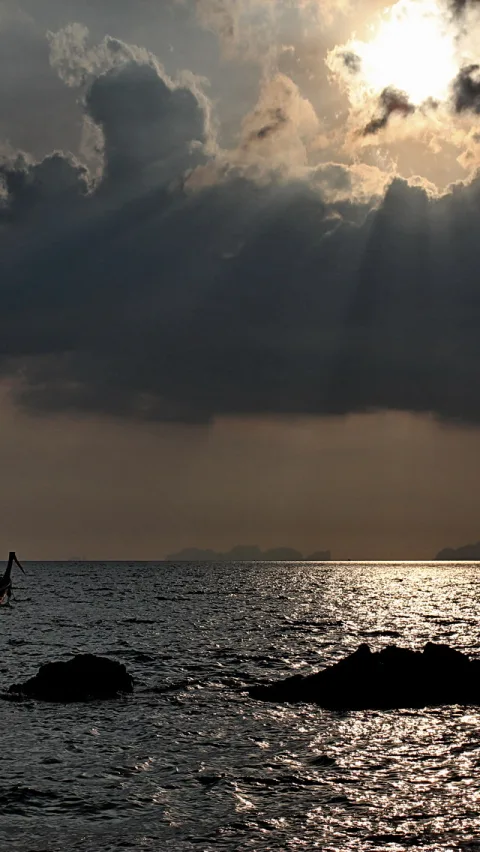 Dunkle Wolken lassen Sonnenstrahlen auf eine Meeresfläche fallen, im Vordergrund ragen dunkle Felsen aus dem Wasser, links ist der Bug eines Bootes zu sehen.
