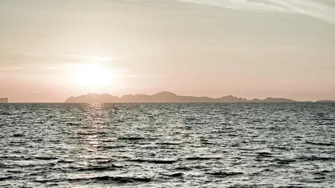 Sonne tief über dem Meer, davor eine dunkle Silhouette einer Inselkette unter einem hellen, rosagrauen Himmel. Die Wasseroberfläche spiegelt das Sonnenlicht in einer hellen Bahn.