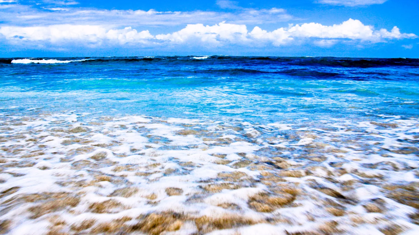 Türkisblaues Meer mit weißer Gischt auf einem flachen, felsigen Ufer, darüber ein blauer Himmel mit weißen Wolken.
