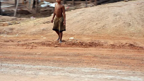 Ein shirtloser Junge in Shorts geht barfuß über eine rote Erdstraße, im Hintergrund stehen einfache Holzbauten und Bambusstangen.