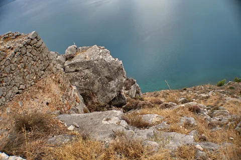 Blick von einer Felsklippe hinab auf graues Gestein, trockenes Gras und eine alte Steinmauer links, dahinter glatte blaugrüne Meeresfläche.