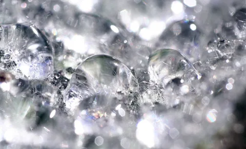 Makroaufnahme von klaren Wassertropfen und Blasen auf einer glitzernden, eisig wirkenden Oberfläche, mit vielen Lichtreflexen und unscharfem Bokeh im Hintergrund.