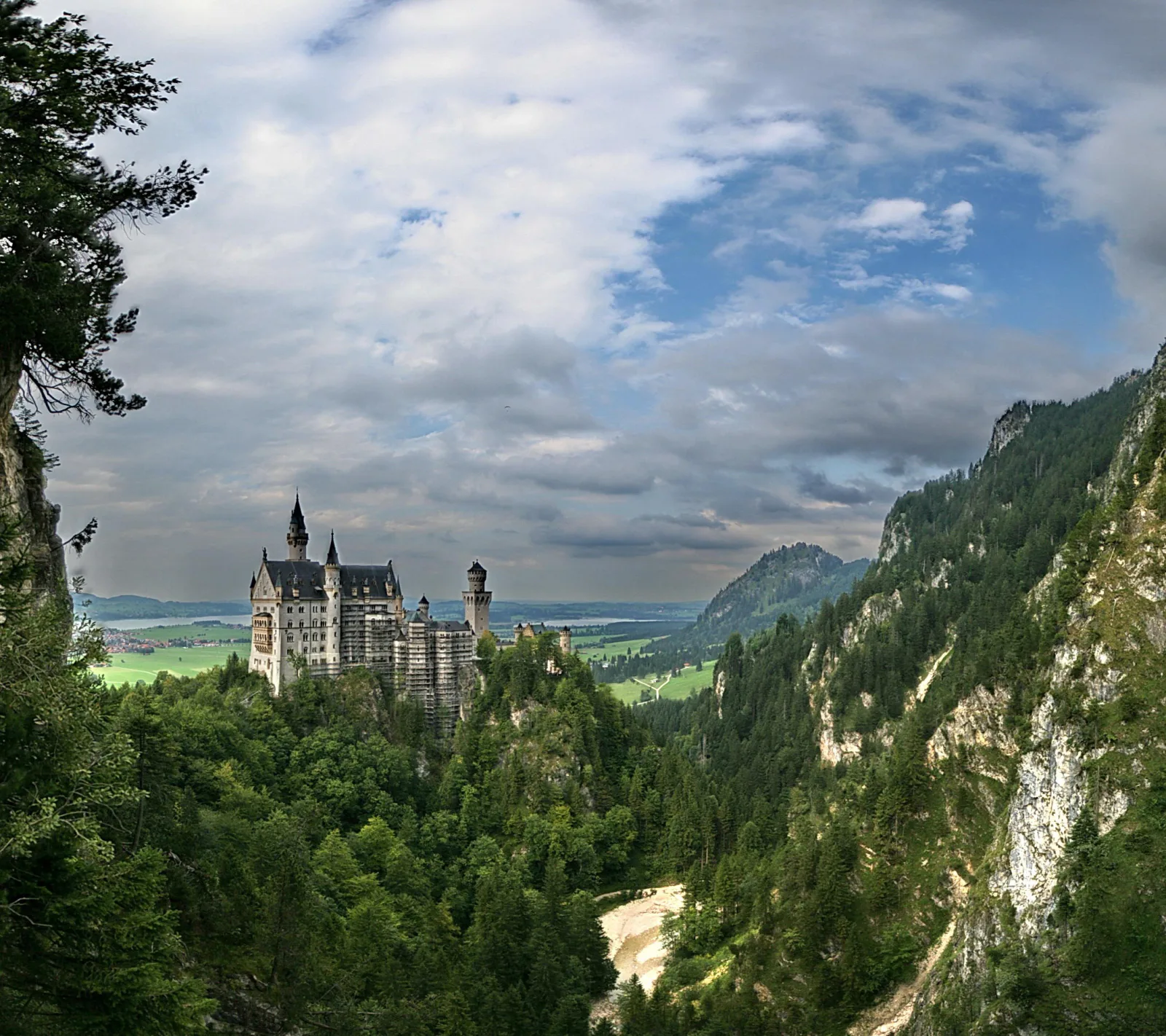Schloss Neuschwanstein auf einem bewaldeten Felsvorsprung, dahinter ein weites Tal mit Bergen und bewölktem Himmel
