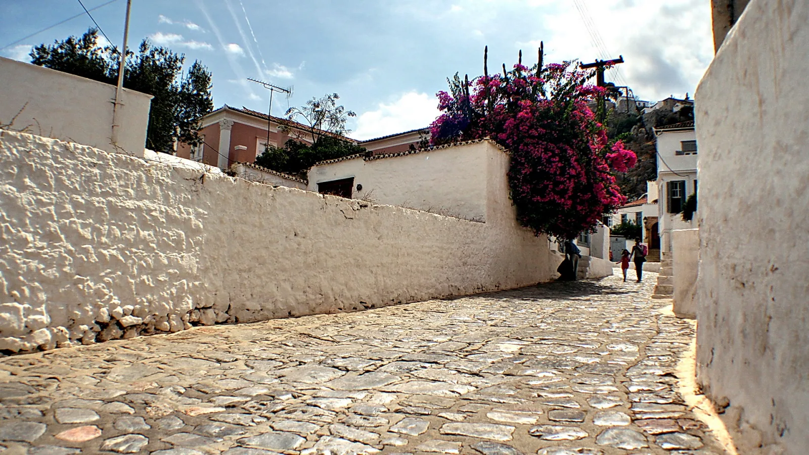 Schmale, kopfsteingepflasterte Gasse zwischen weiß getünchten Mauern. Rechts wächst pinkfarbene Bougainvillea über eine Mauer, im Hintergrund gehen zwei Personen die Gasse hinauf.