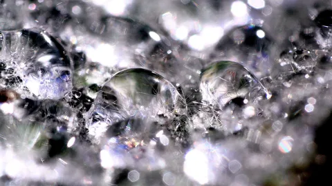 Makroaufnahme vieler glänzender Wasserblasen und Tropfen auf einer grauen, glitzernden Oberfläche, mit hellen Lichtreflexen.