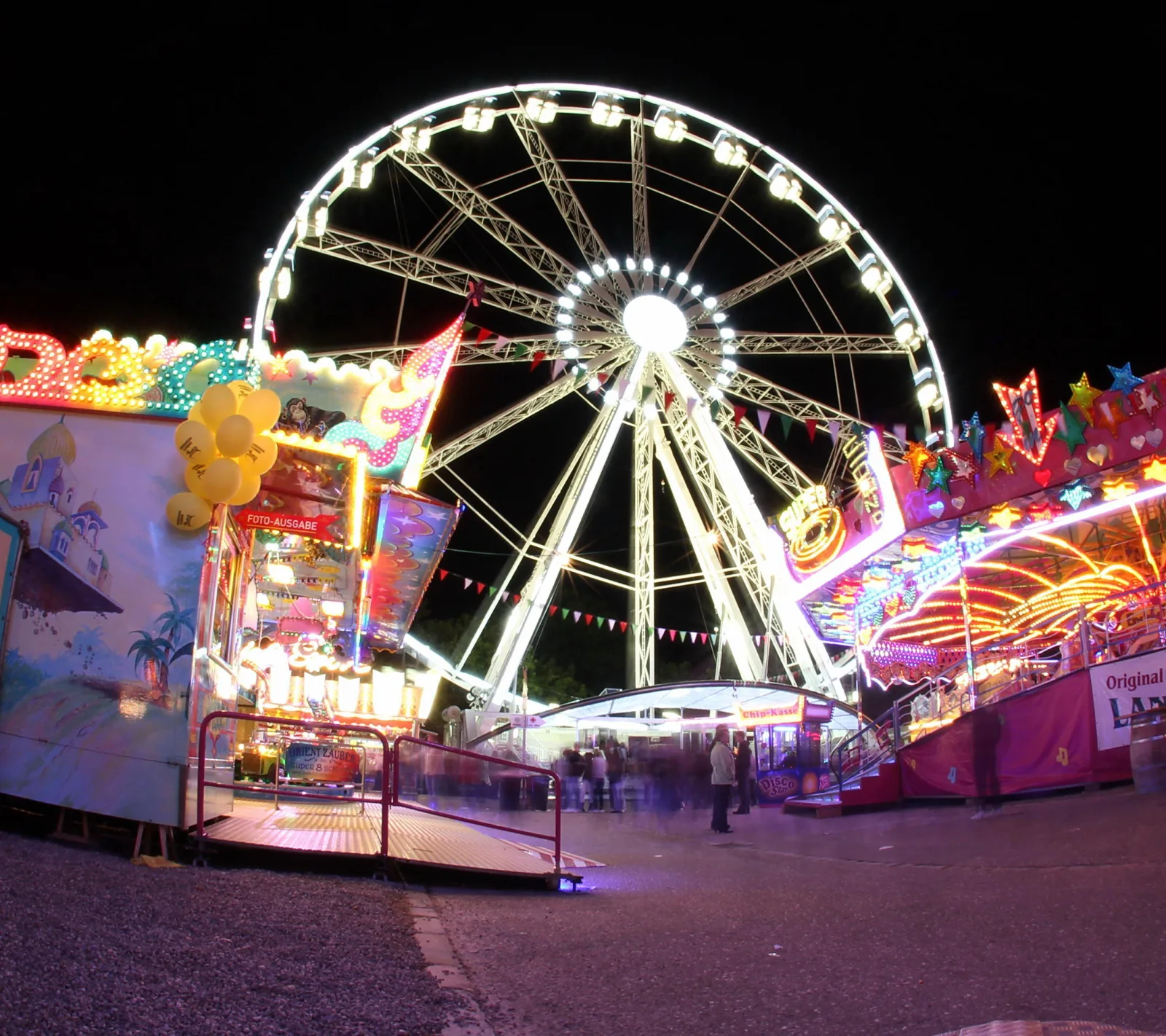 Beleuchtetes Riesenrad vor dunklem Nachthimmel, davor Fahrgeschäfte und Buden mit bunten Lichtern und verschwommenen Passanten.