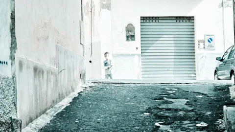 Ein kleiner Junge steht in einer hellen, verwitterten Gasse vor einem geschlossenen Rolltor, rechts parkt ein silbernes Auto.