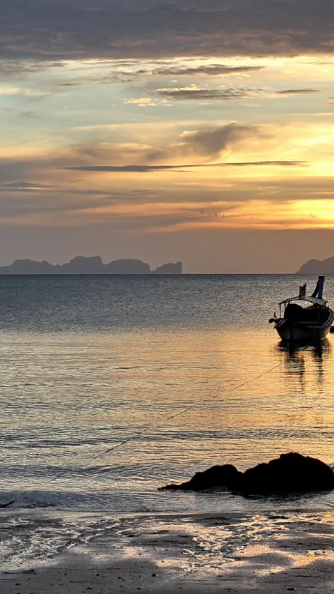 Ein kleines Holzboot liegt rechts im ruhigen Meer, im Hintergrund zeichnen sich Felsinseln vor einem orangefarbenen Abendhimmel ab, im Vordergrund liegen dunkle Felsen am nassen Sandstrand.
