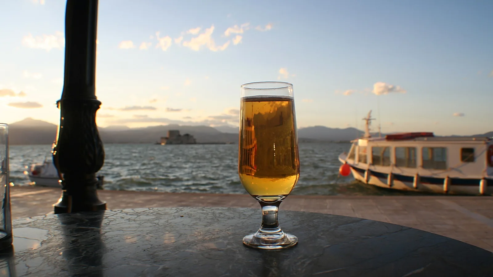 Ein Glas helles Bier steht auf einem dunklen Marmortisch. Im Hintergrund liegen Wasser, eine kleine Festung auf einer Insel, Berge und ein weißes Ausflugsboot unter einem abendlichen Himmel.