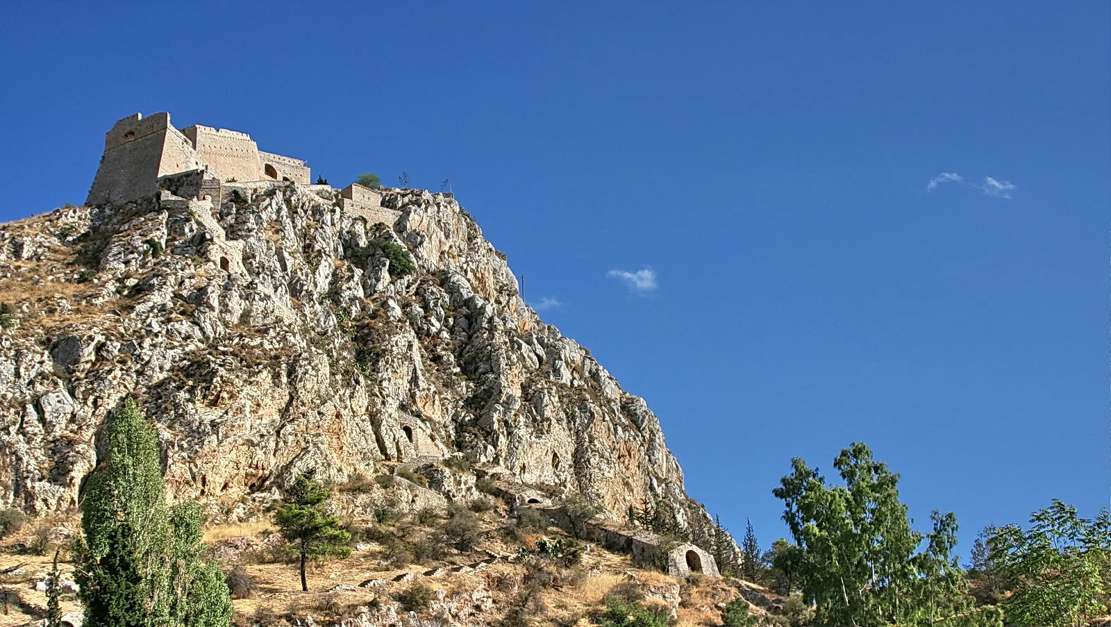 Steinerne Festung mit Mauern und Türmen auf einem felsigen Hügel vor blauem Himmel, darunter Bäume und trockenes Gras.