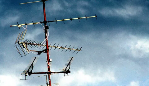 Mehrere Fernsehantennen an einem rot-weißen Mast vor dunkel bewölktem blauem Himmel.