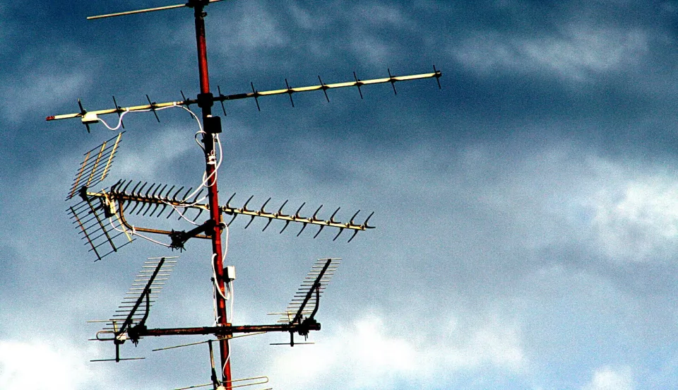 Mehrere Fernsehantennen an einem rot-weißen Mast vor dunkel bewölktem blauem Himmel.