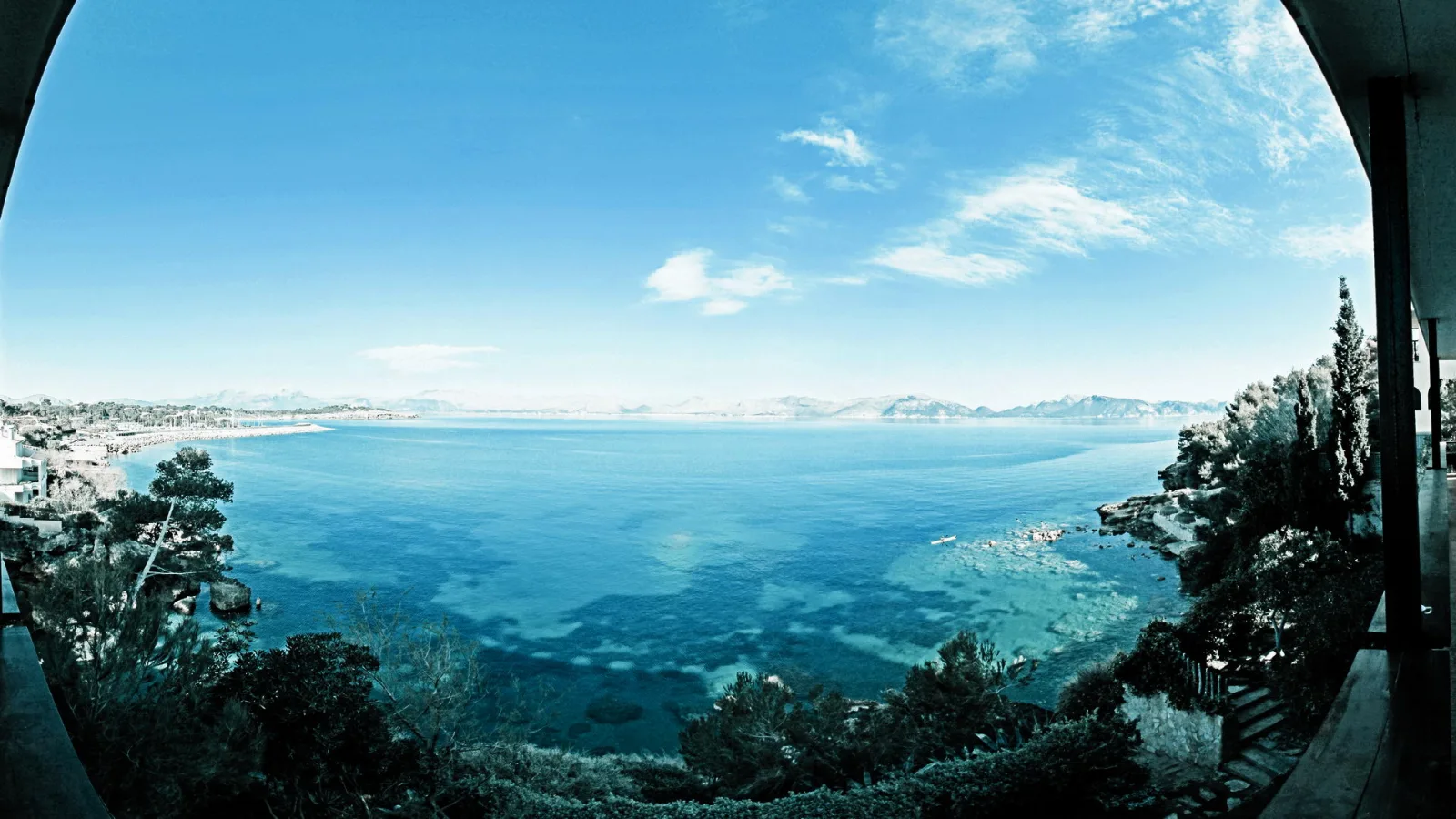 Panoramablick von einer Terrasse auf eine türkisfarbene Meeresbucht mit felsiger Küste, Sträuchern im Vordergrund und blauem Himmel mit Wolken; am Horizont Bergketten und ein heller Küstenstreifen.