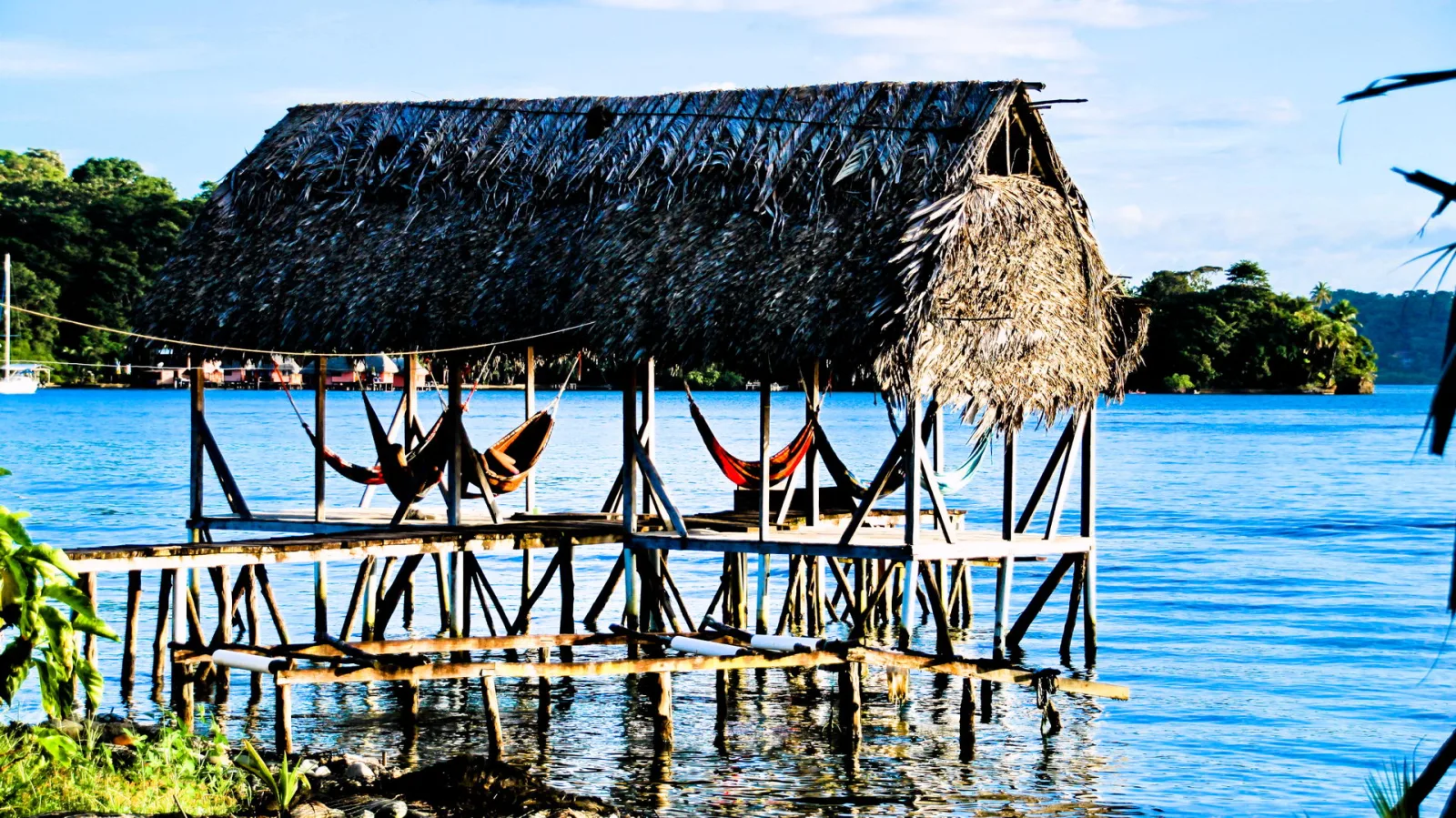 Ein Holzsteg mit strohgedecktem Dach steht auf Stelzen im blauen Wasser. Darunter hängen mehrere Hängematten, im Hintergrund sind grüne Inseln und ein weißes Segelboot zu sehen.