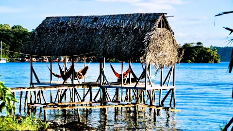 Ein Holzsteg mit strohgedecktem Dach steht auf Stelzen im blauen Wasser. Darunter hängen mehrere Hängematten, im Hintergrund sind grüne Inseln und ein weißes Segelboot zu sehen.
