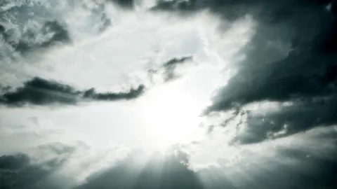 Dichte Wolkendecke mit einer hellen Sonne in der Mitte, von der Lichtstrahlen nach unten fallen; rechts dunkle Gewitterwolken, links helleres Grau.