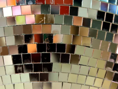 Nahaufnahme einer gewölbten Fläche aus kleinen quadratischen Spiegelkacheln in Gold-, Rot-, Grün- und Grautönen, in denen sich die Umgebung spiegelt.