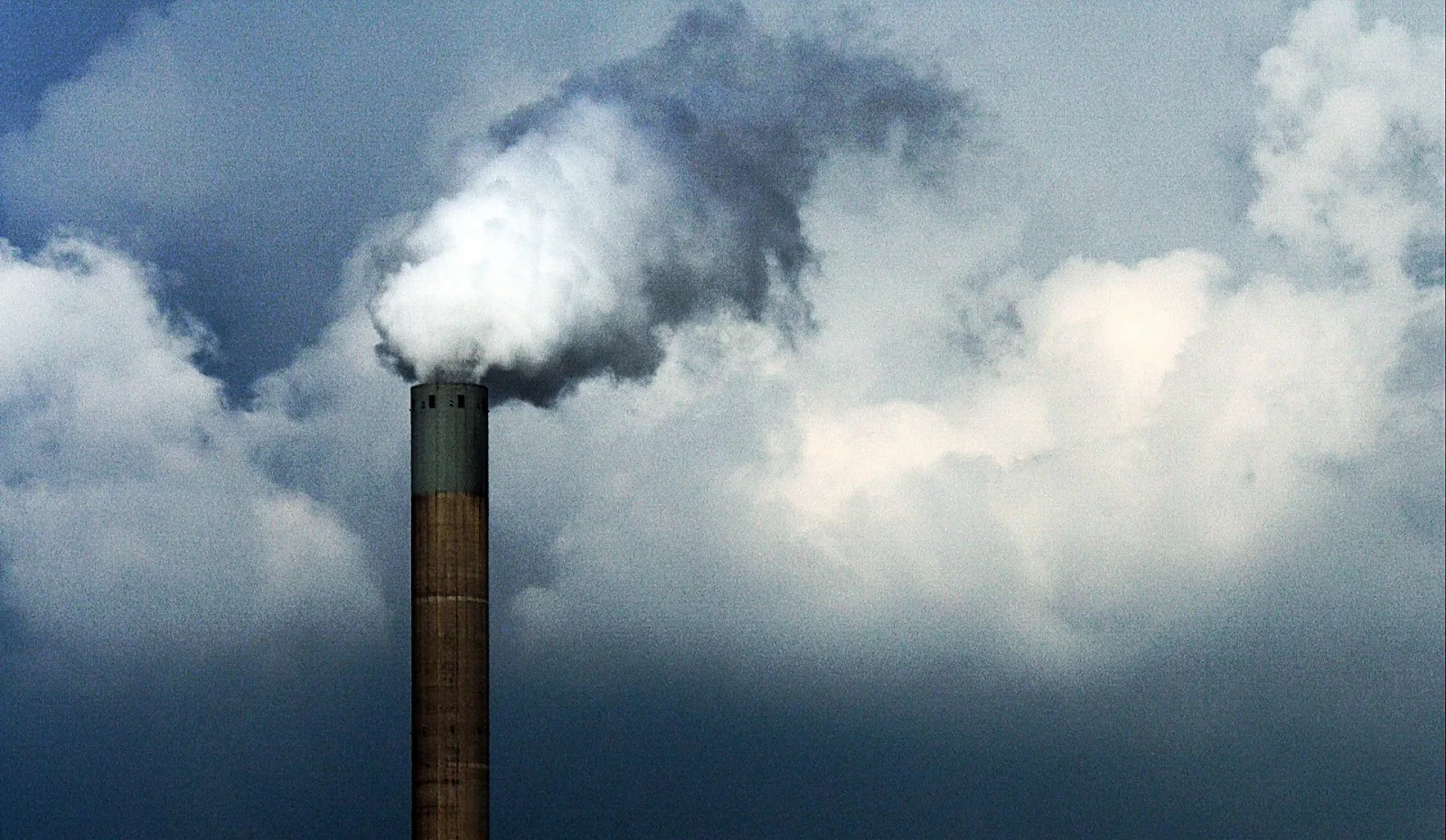 Ein schlanker Industrieschornstein aus Backstein mit grünlicher Kappe ragt ins Bild, aus dem eine weiße bis graue Rauchfahne in einen bewölkten, blaugrauen Himmel steigt.