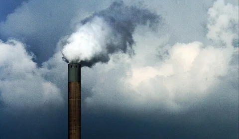Ein schlanker Industrieschornstein aus Backstein mit grünlicher Kappe ragt ins Bild, aus dem eine weiße bis graue Rauchfahne in einen bewölkten, blaugrauen Himmel steigt.