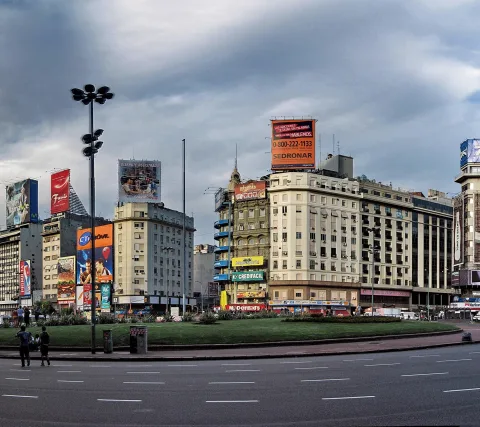 Stadtansicht mit Kreisverkehr, grüner Mittelinsel und Hochhäusern mit Werbetafeln unter bewölktem Himmel in Buenos Aires.