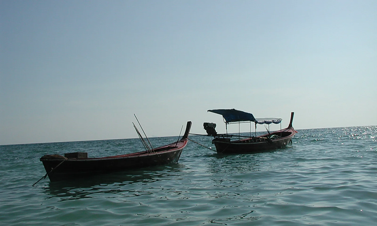Zwei hölzerne Longtailboote mit roten Rümpfen liegen auf ruhigem, türkisfarbenem Meer unter dunstigem Himmel.