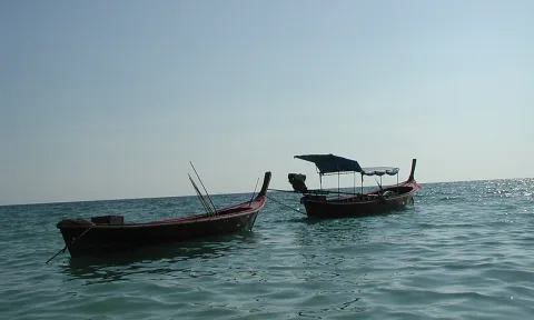 Zwei hölzerne Longtailboote mit roten Rümpfen liegen auf ruhigem, türkisfarbenem Meer unter dunstigem Himmel.