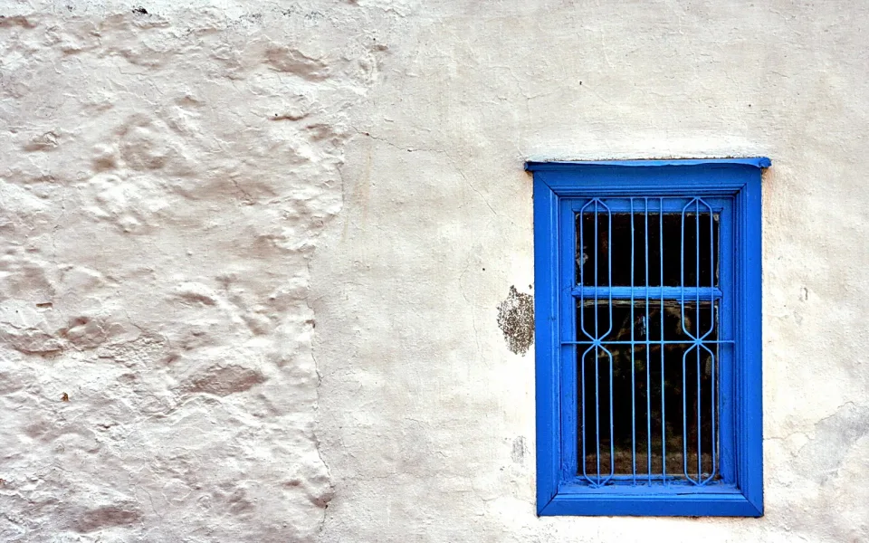 Ein rechteckiges Fenster mit leuchtend blauem Rahmen und blauen Metallstäben sitzt rechts in einer weiß verputzten, unebenen Hauswand.