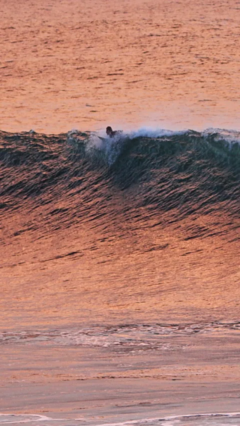 Ein Surfer liegt auf dem Brett und gleitet in einer dunklen Welle, die sich vor einer orangefarbenen Meeresfläche bei Sonnenuntergang aufbaut.
