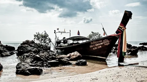 Ein dunkles Longtailboot liegt auf einem flachen Sandstrand neben dunklen Felsen, am Heck hängen bunte Stoffbänder, darüber ein bewölkter Himmel.