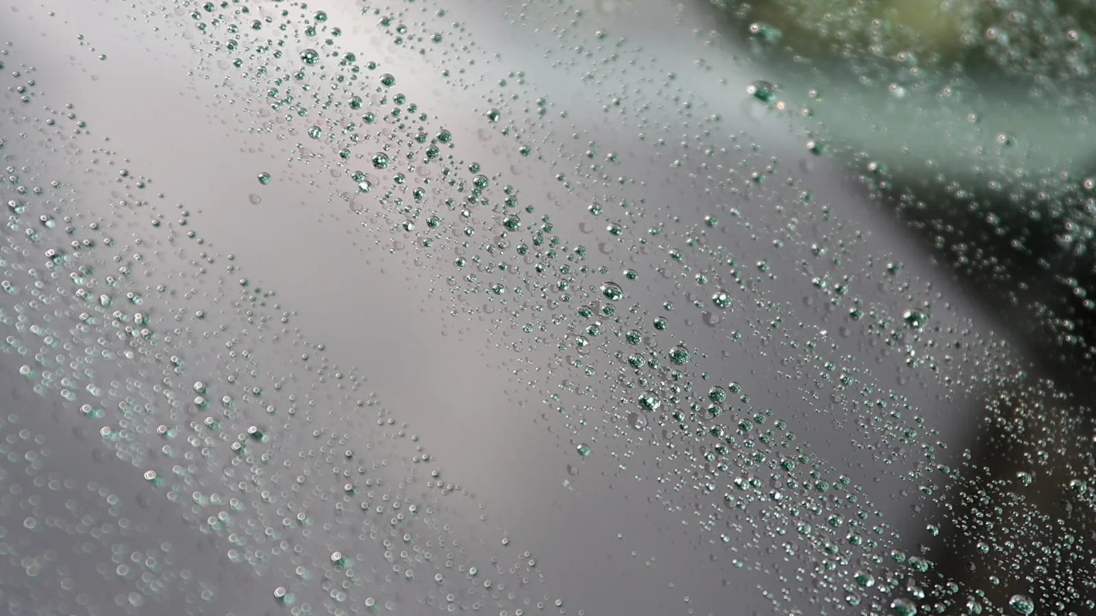 Viele kleine Wassertropfen perlen diagonal über eine graue, glatte Glasscheibe, rechts unscharf grünlicher Hintergrund.