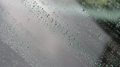 Viele kleine Wassertropfen perlen diagonal über eine graue, glatte Glasscheibe, rechts unscharf grünlicher Hintergrund.