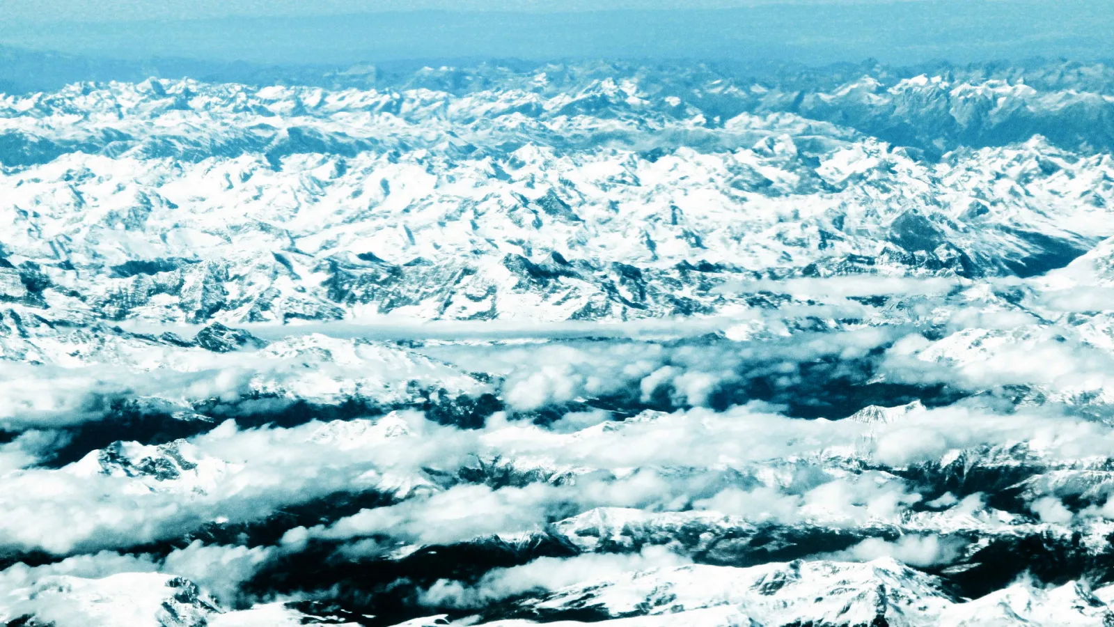 Luftaufnahme einer schneebedeckten Gebirgskette, zwischen den Gipfeln liegen Wolkenbänder, der obere Bildrand ist blau und diesig.