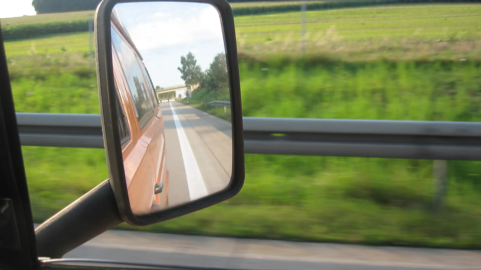 Blick durch das Fenster auf einen Seitenspiegel, der einen orangefarbenen Transporter und eine Landstraße mit weißer Mittellinie zeigt, dahinter grüne Felder und eine Leitplanke.