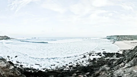 Breite Bucht mit weißer Brandung, dunklen Felsen im Vordergrund, einem Sandstrand rechts und einer steilen Felsküste am Horizont unter bedecktem Himmel.