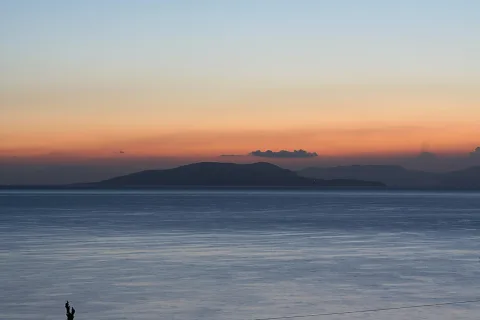 Ruhiges Meer unter einem orangeroten Abendhimmel, im Hintergrund dunkle Inseln und Hügelketten, am unteren Rand zwei kleine Silhouetten auf einem Pfahl.